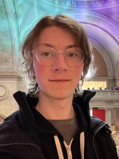 Me standing in The Met building in NYC on a day I went to th emuseum. I'm wearing a light jacket and hooded jumper, and smiling.