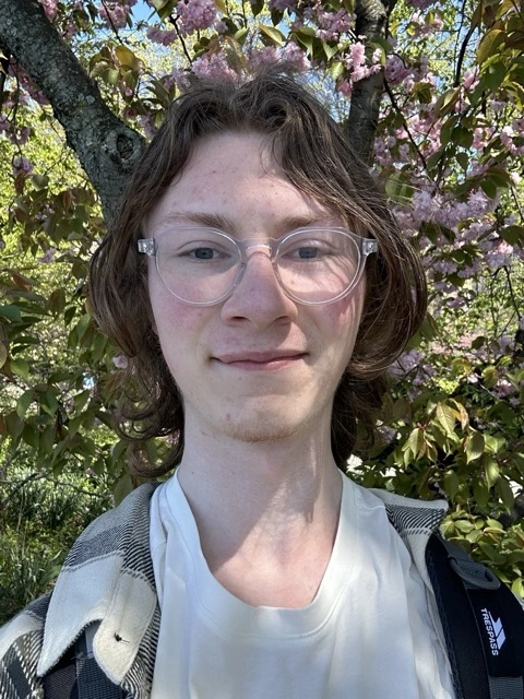 Me standing in front of cherry blossom trees in spring, smiling, wearing a light coloured shirt and jumper.