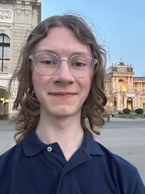 Me, with long hear wearing a navy blue polo shirt, in Vienna, standing in front of a museum, smiling with joy.