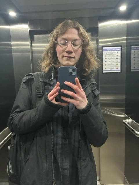 Me standing in an elevator with a mirror, wearing a black coat and a navy jumper, taking a selfie.